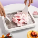 Bright blue rectangular rolled ice cream maker pan with two plastic scrapers rolling up fresh strawberry ice cream on a kitchen table.