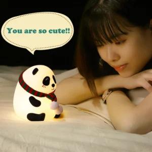 Cute panda silicone night light glowing with a soft warm light on a wooden bedside table in a dark cozy bedroom.